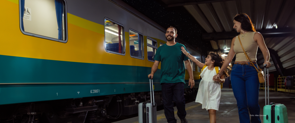 Família com pai, mãe e filha sorrindo, com malas, caminhando ao lado do trem de passageiros da Vale (amarelo e verde) à noite em uma plataforma de estação.